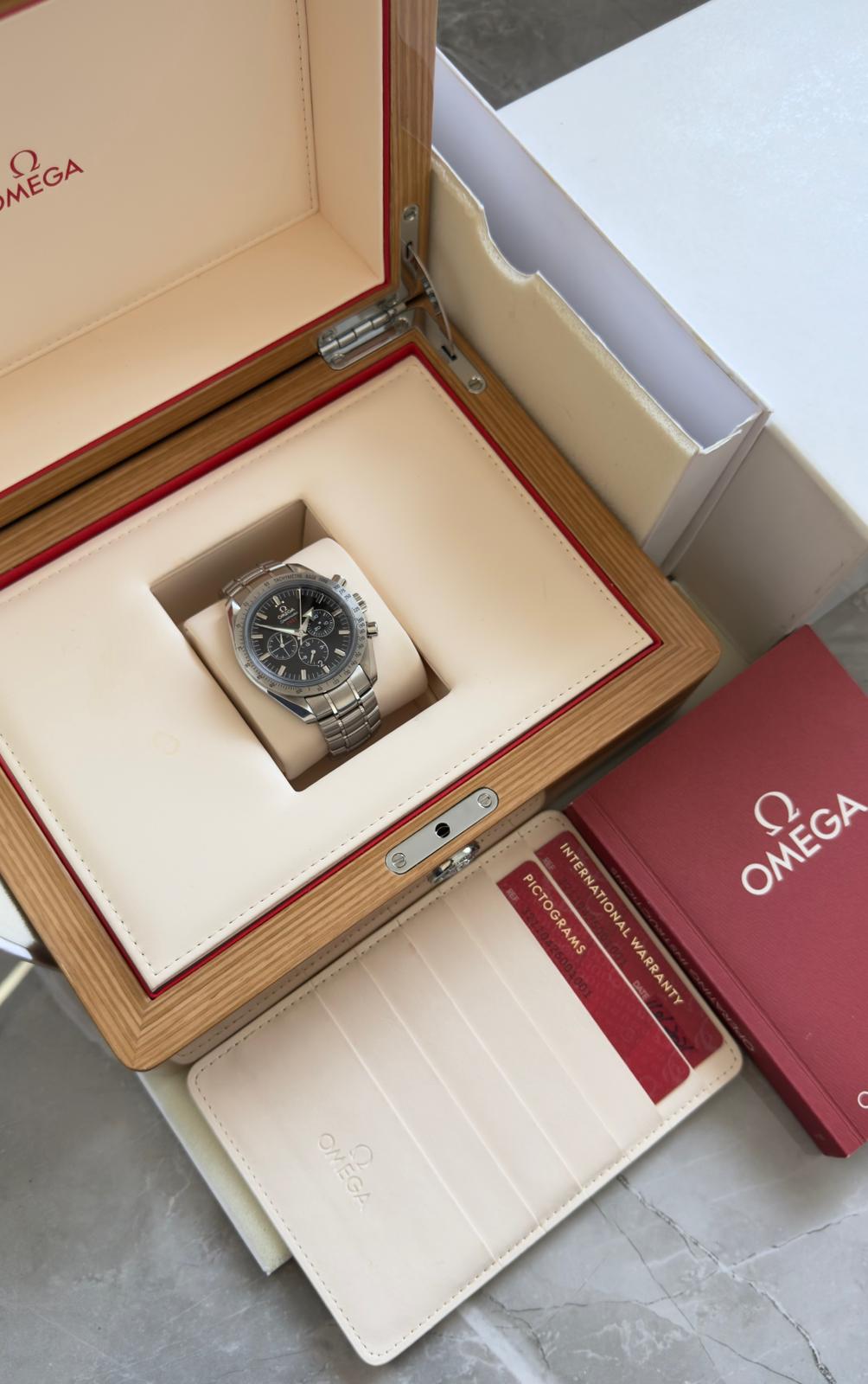 Omega Speedmaster Broad Arrow displayed in an open wood-tone box with cream interior, red booklet and cards in a document holder.