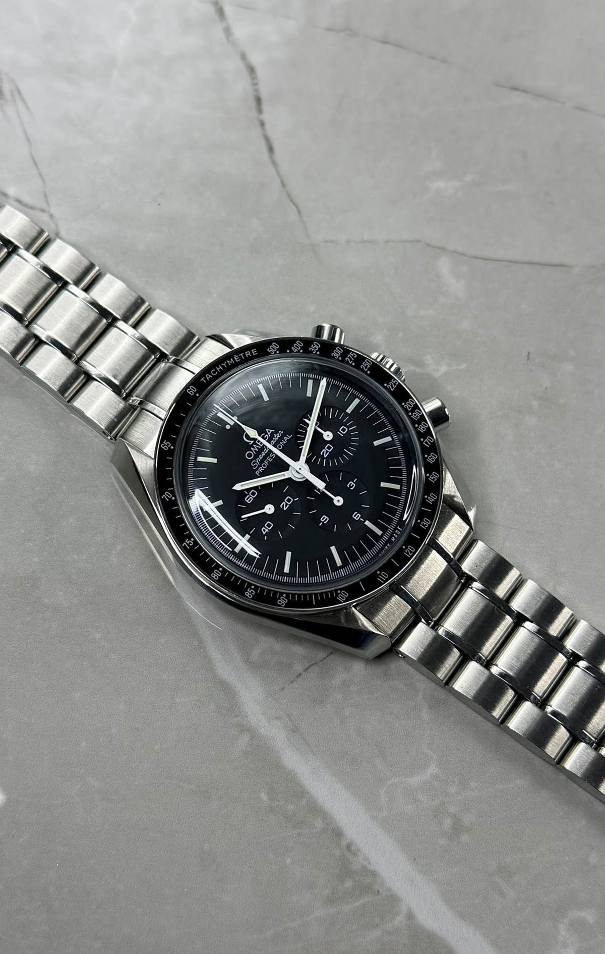 Omega Speedmaster with a black chronograph dial and bracelet laid diagonally on a grey surface.