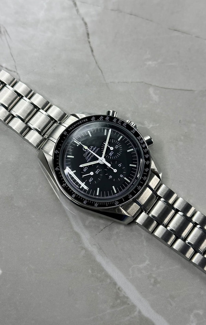 Omega Speedmaster with a black chronograph dial and bracelet laid diagonally on a grey surface.