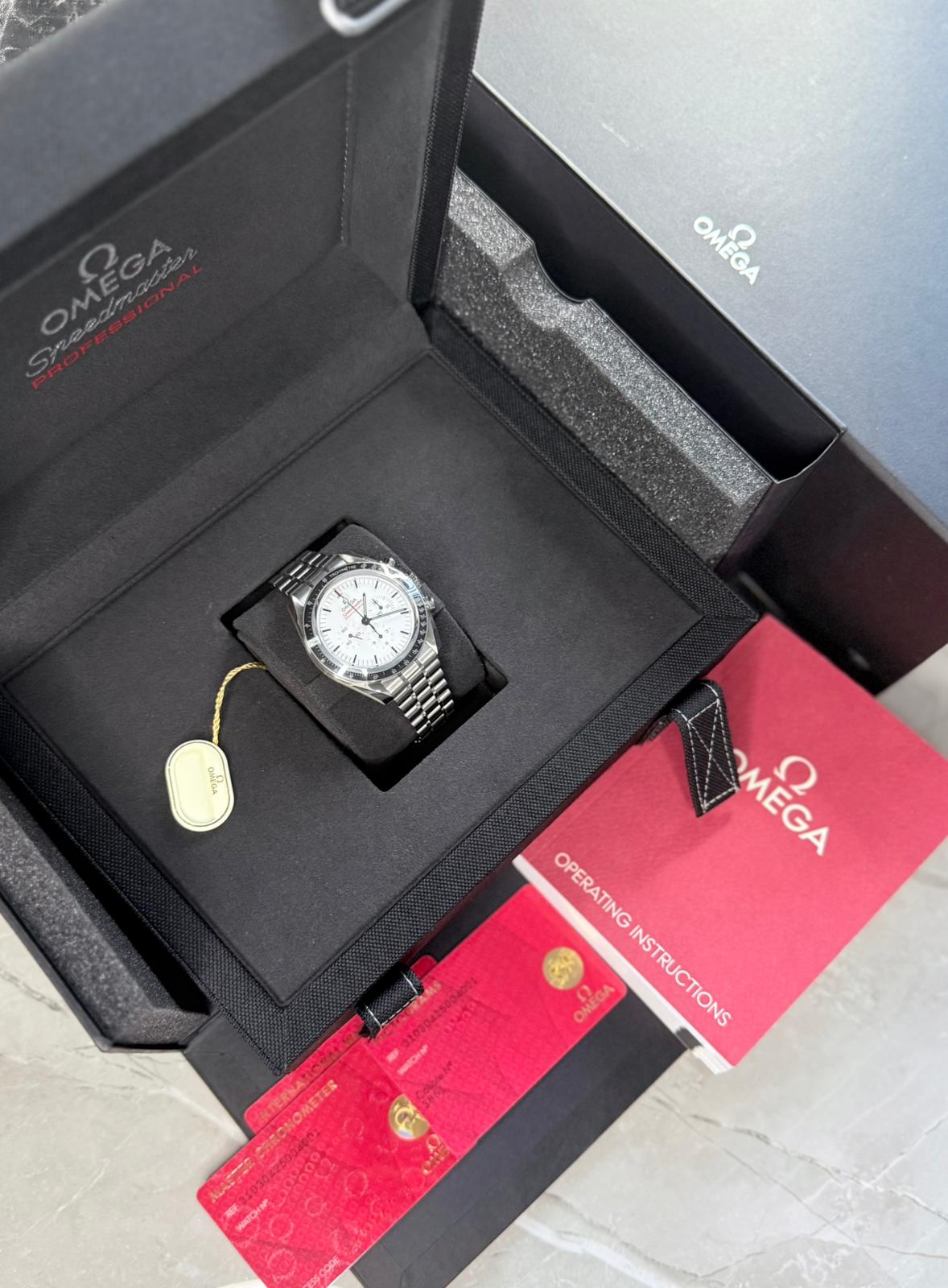 Omega Speedmaster displayed in an open black presentation box with white dial, tag, red cards and printed booklet.