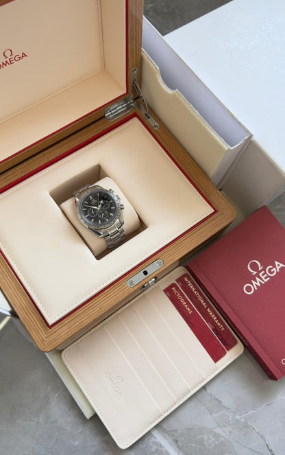 Omega Speedmaster Broad Arrow displayed in an open wood-tone box with cream interior, red booklet and cards in a document holder.